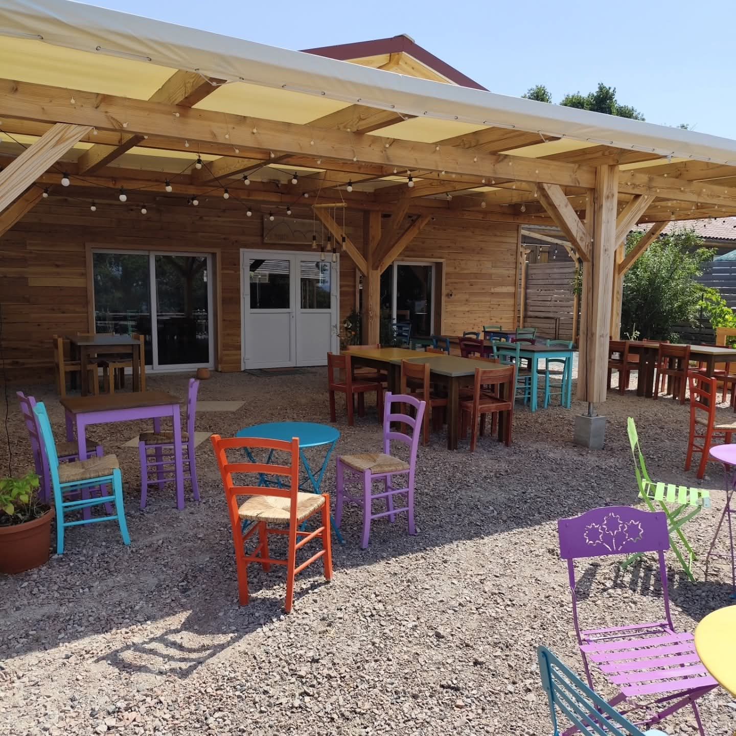 Café en plein air avec terrasse en bois, chaises colorées et tables sur gravier sous un auvent.