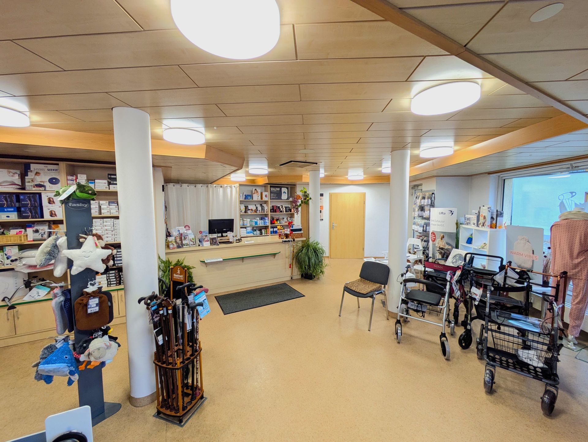 Apothekerin steht hinter einem hellen Holzthekenbereich in einer Apotheke mit Regalen voller Arzneimittel und Pflegeprodukte.