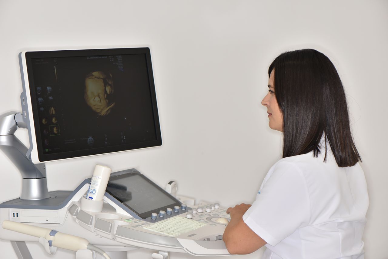 Mujer con uniforme blanco operando una máquina de ultrasonido, mirando un monitor con una imagen.