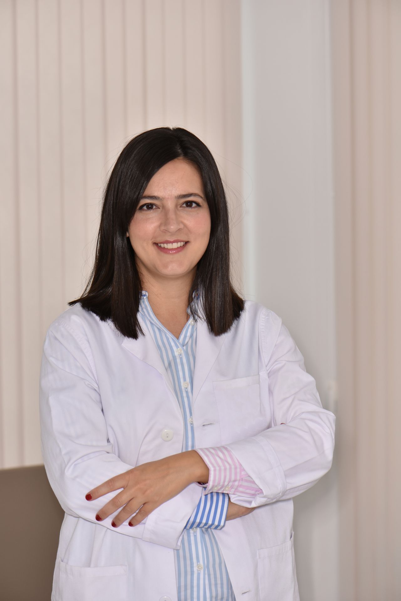 Mujer con bata blanca, sonriendo, con los brazos cruzados. De pie en una habitación bien iluminada.