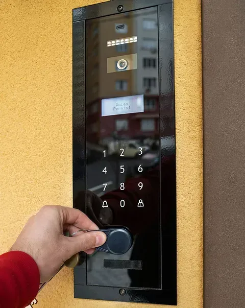 Mano sosteniendo un llavero sobre un teclado de intercomunicador negro con teclado numérico y pantalla, montado en una pared amarilla.
