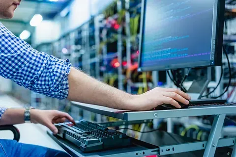 Hombre usando una computadora en una sala de servidores, monitoreando datos, camisa a cuadros azul y blanca.