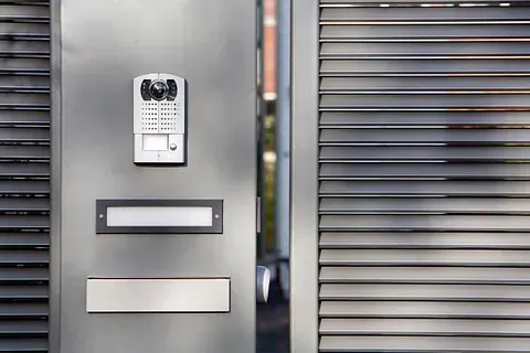 Puerta de metal gris con timbre con cámara, buzón y placa de dirección.
