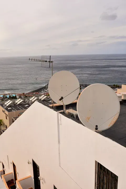 Dos antenas parabólicas blancas en el tejado de un edificio blanco, con el océano al fondo bajo un cielo nublado.