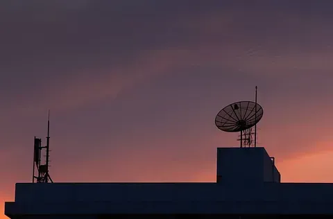 Antenas silueteadas y antena parabólica encima de un edificio contra un cielo colorido al atardecer.