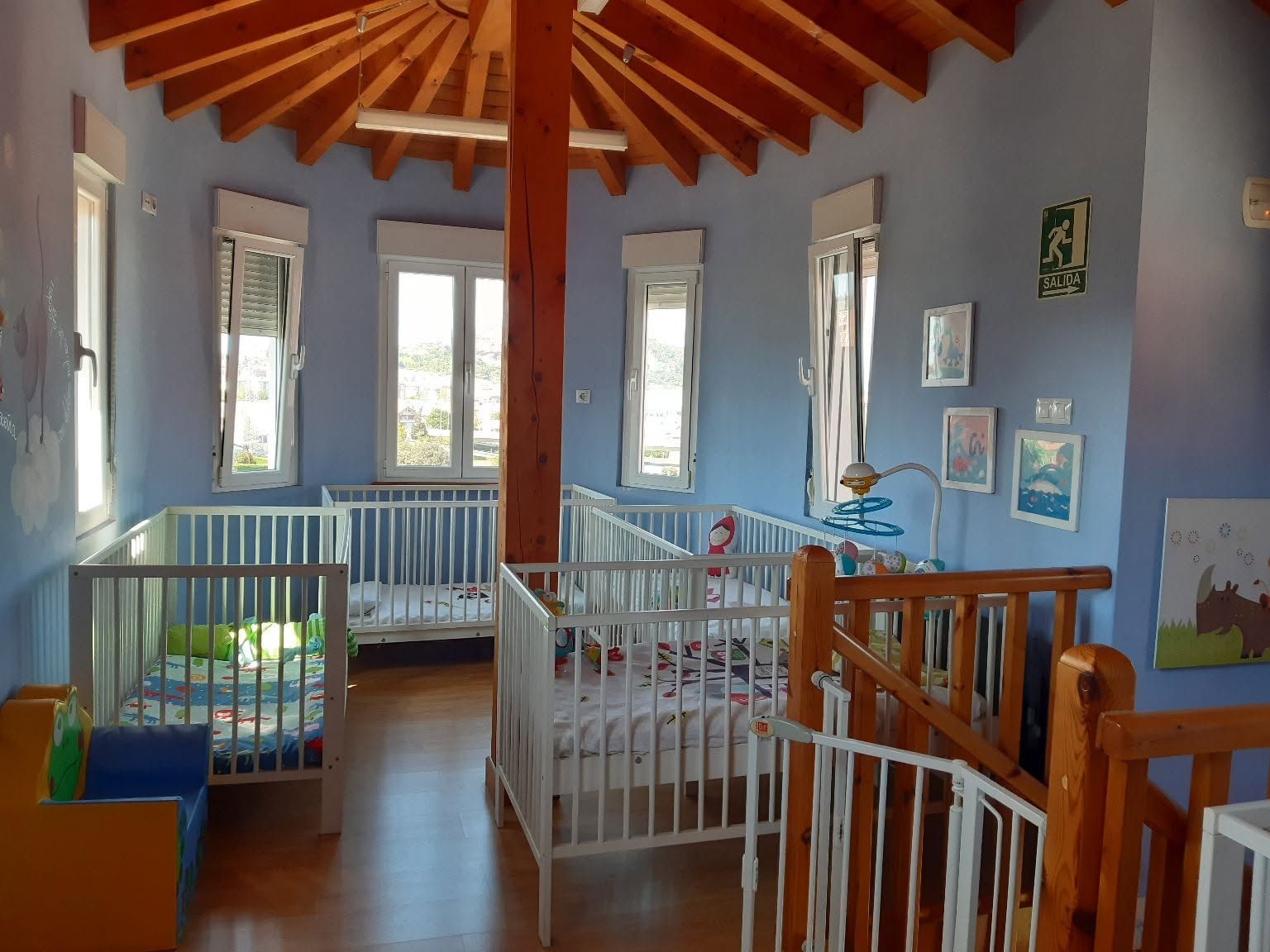 Habitación infantil luminosa con varias cunas blancas, paredes azules y techo con vigas de madera.