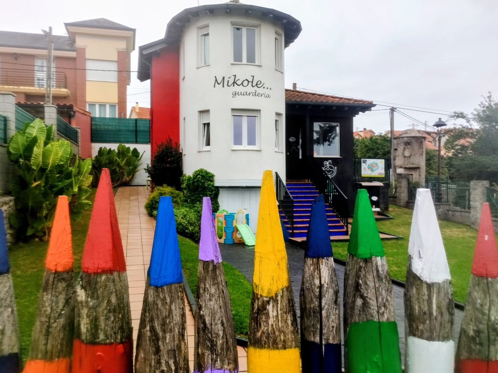 Valla colorida con forma de lápiz frente a un edificio cilíndrico blanco con un letrero, junto a un jardín y un patio.