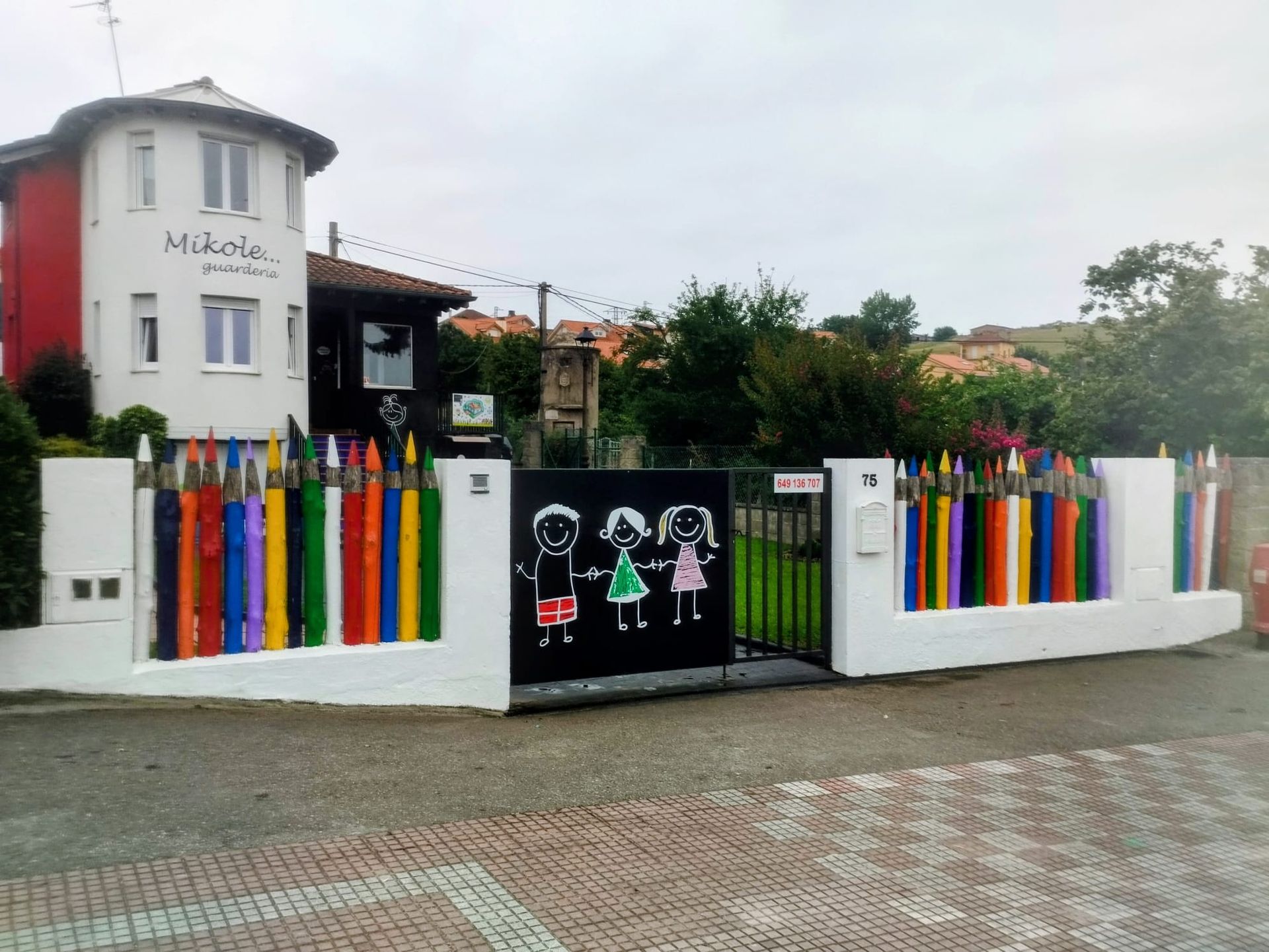 Entrada vallada de colores con listones de arcoíris y una puerta negra con dibujos infantiles junto a un edificio.