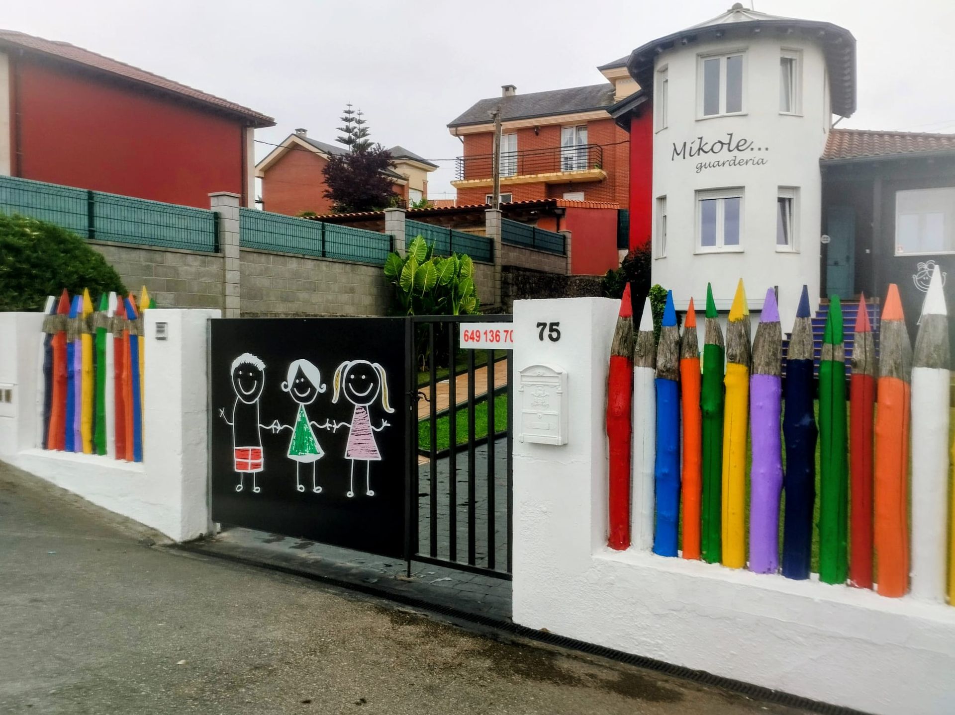 Una colorida valla hecha con lápices frente a una guardería, con dibujos infantiles en la puerta y un edificio blanco detrás.