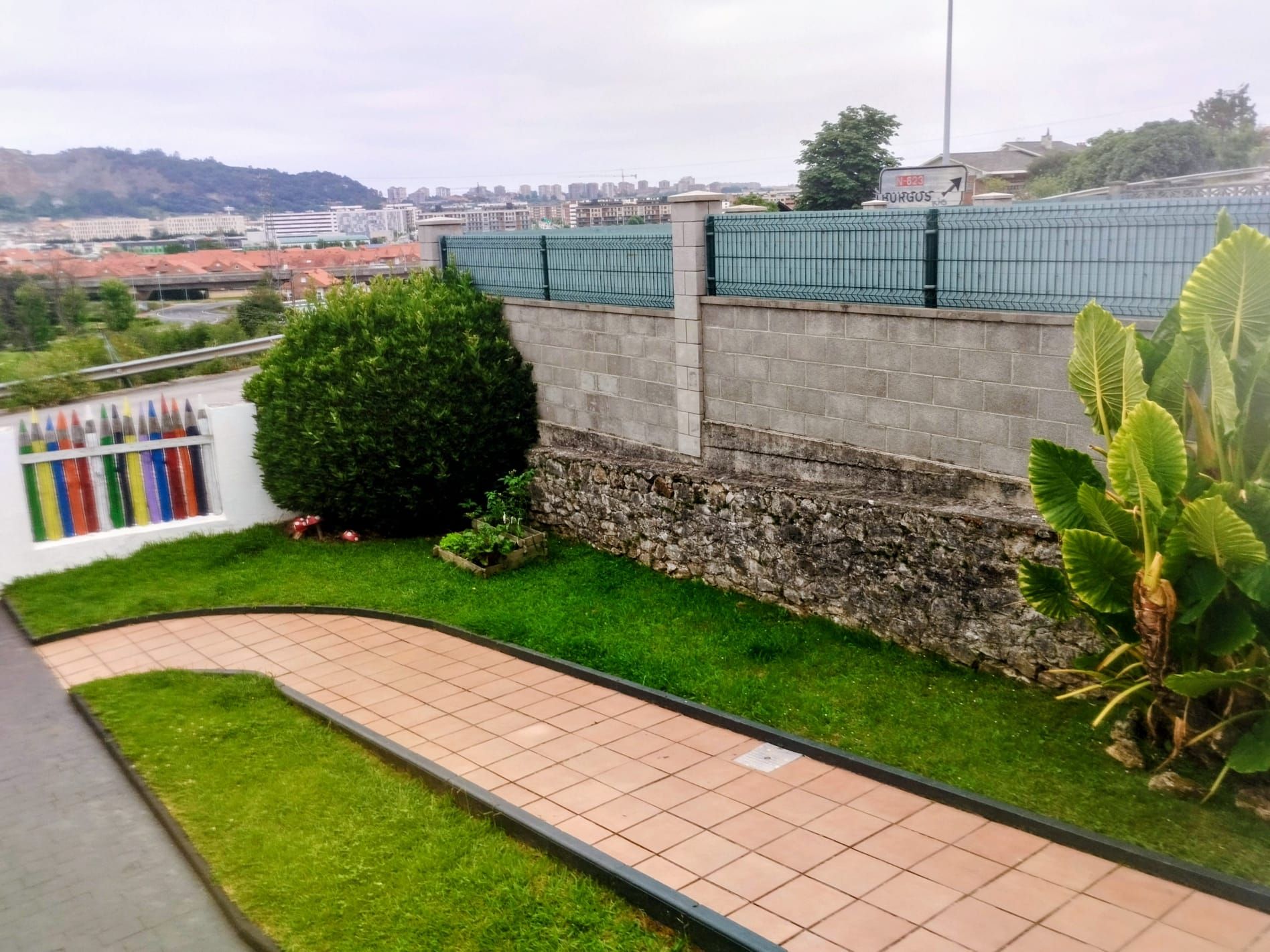 Patio curvo de ladrillo junto a un jardín con césped, arbustos verdes, muro de piedra y valla con vistas a una ladera.