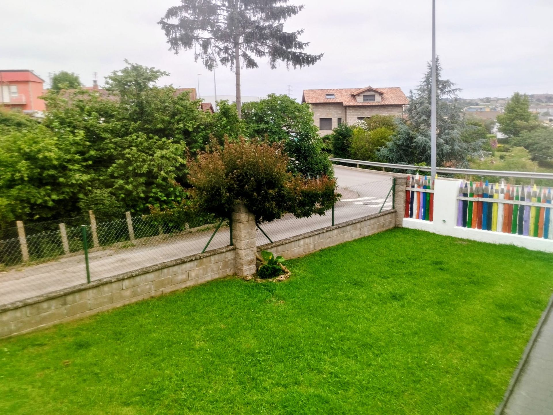 Jardín trasero con un pequeño árbol, valla y barandilla colorida con vistas a las casas y al cielo nublado.