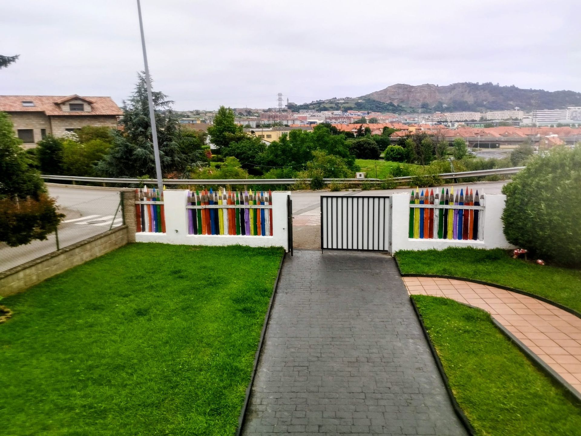Jardín delantero con césped verde, camino pavimentado, puerta blanca y casas en la ladera de la colina a lo lejos.