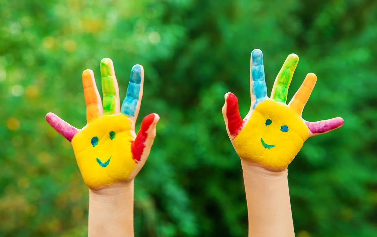 Dos manos pintadas de colores con rostros sonrientes levantadas sobre un fondo verde difuminado.
