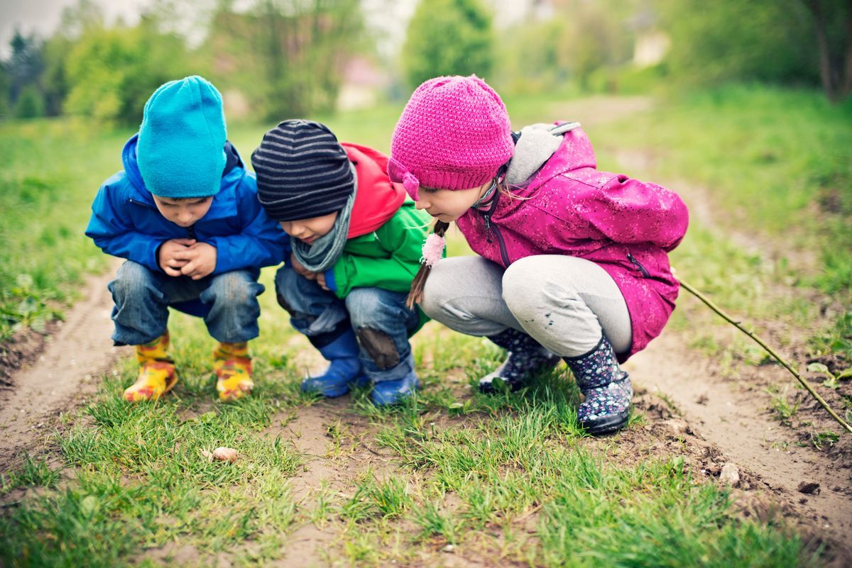 Tres niños con coloridos sombreros se agachan juntos en un sendero cubierto de hierba al aire libre.