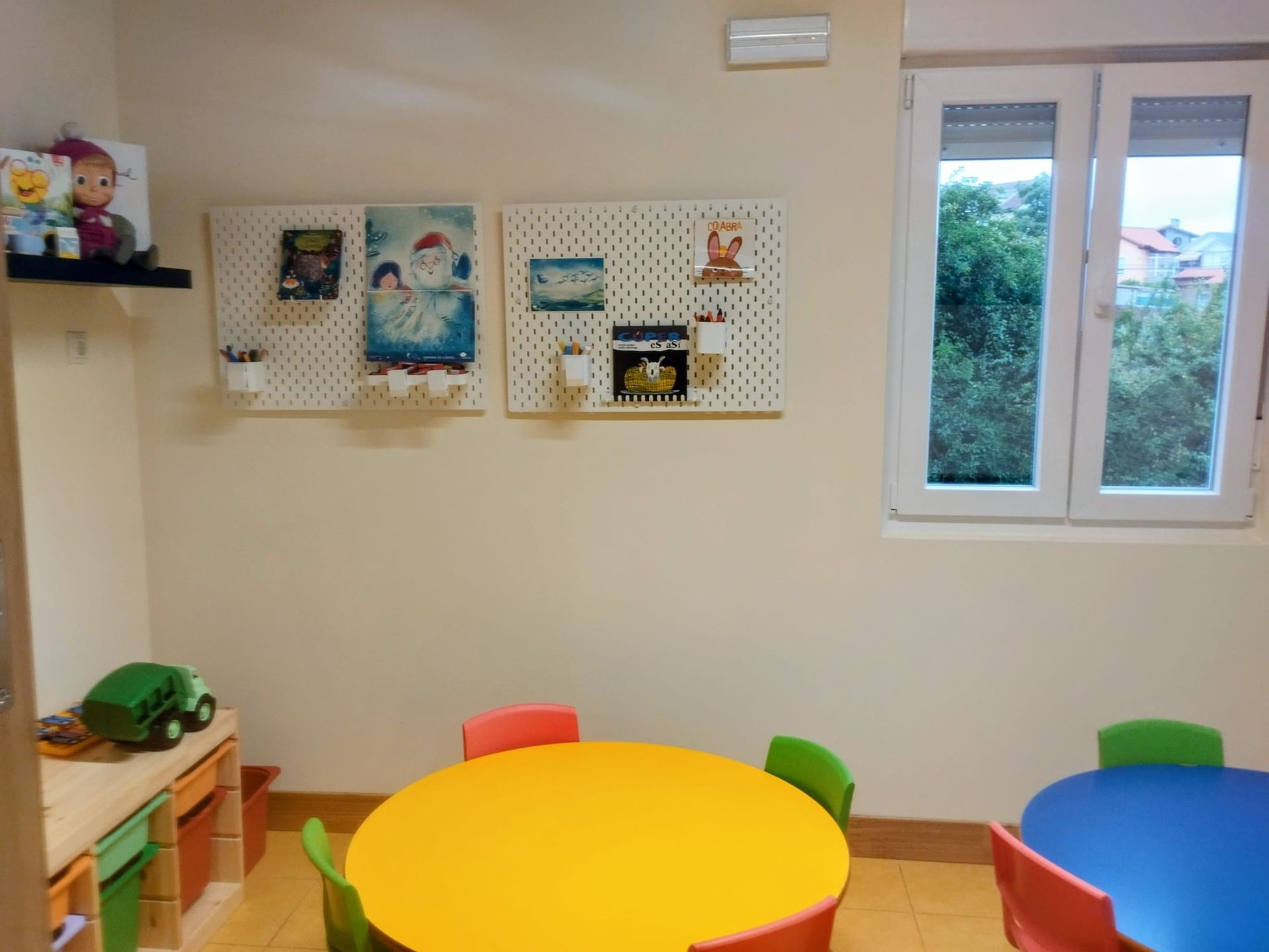 Aula preescolar colorida con mesas pequeñas, sillas, dibujos en las paredes y una ventana que da a los árboles del exterior.