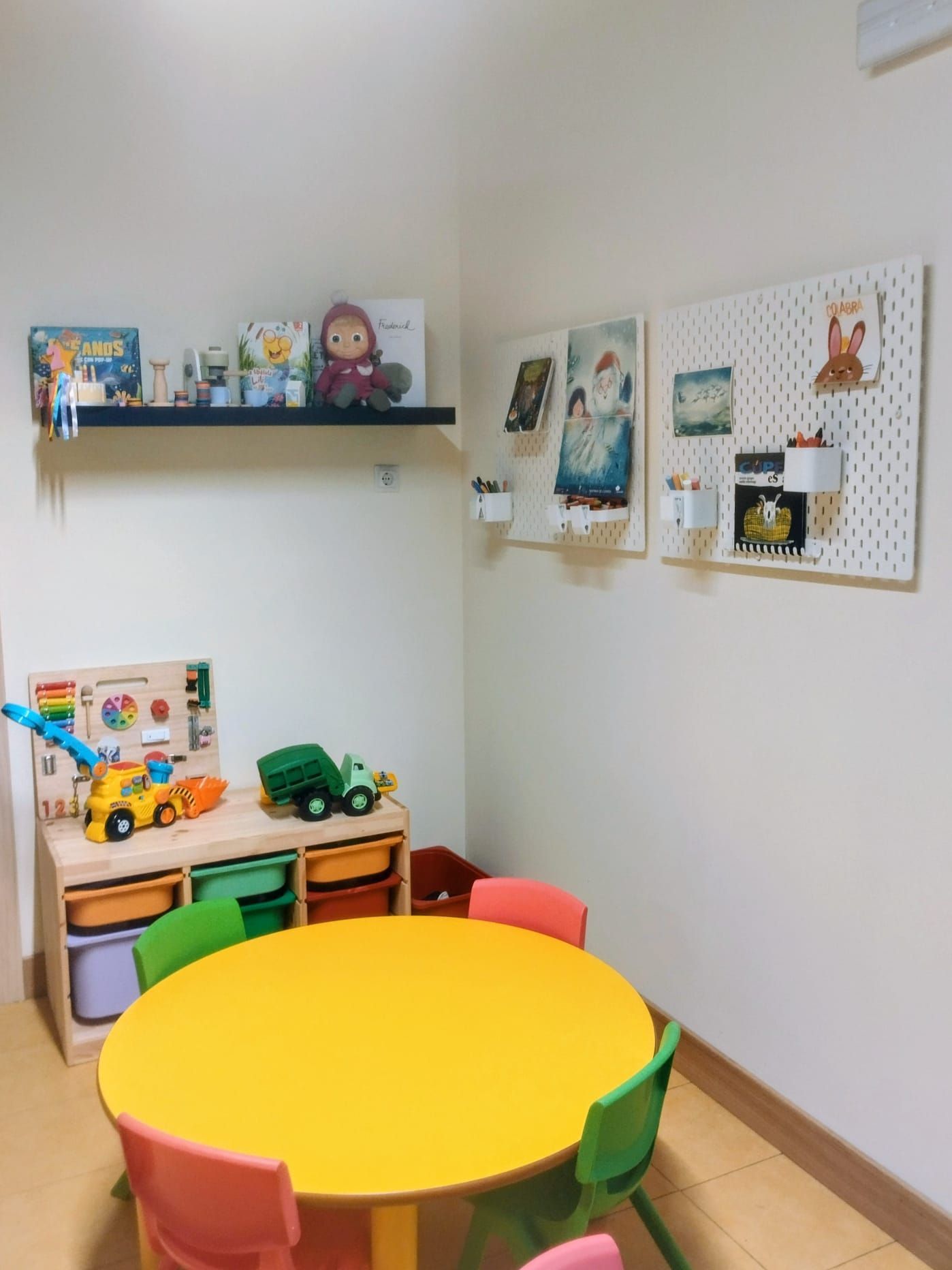 Rincón de juegos infantiles con una mesa amarilla, sillas coloridas, juguetes y obras de arte en las paredes blancas.