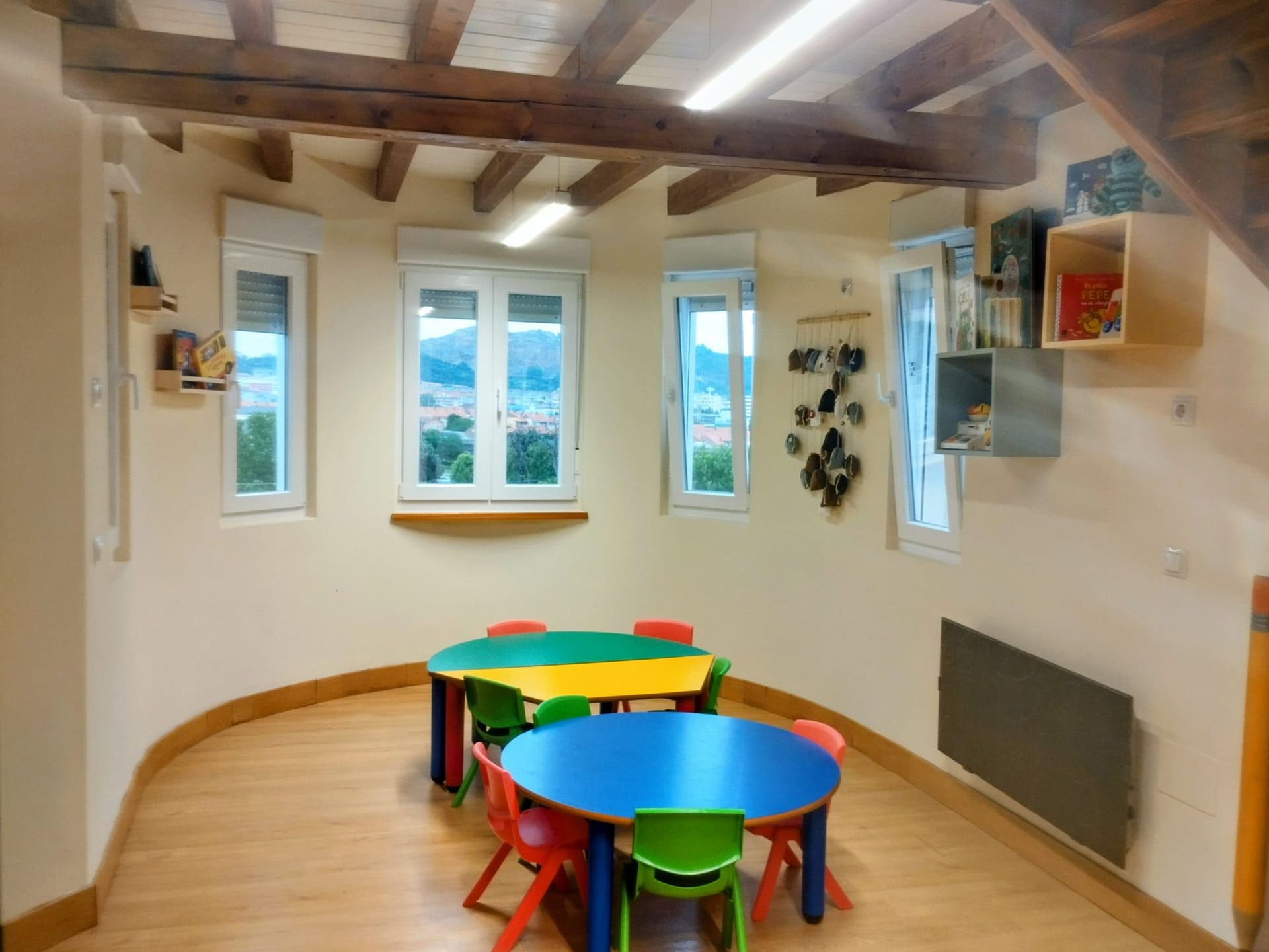 Sala de juegos infantil colorida con mesas redondas, taburetes, ventanas y vigas de madera a la vista.