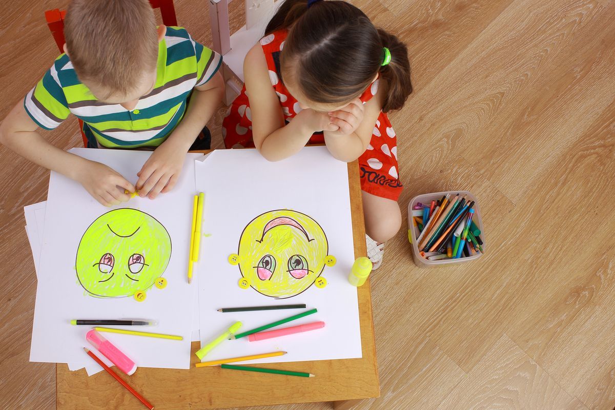 Dos niños coloreando caras de dibujos animados en una mesa con crayones y papel.