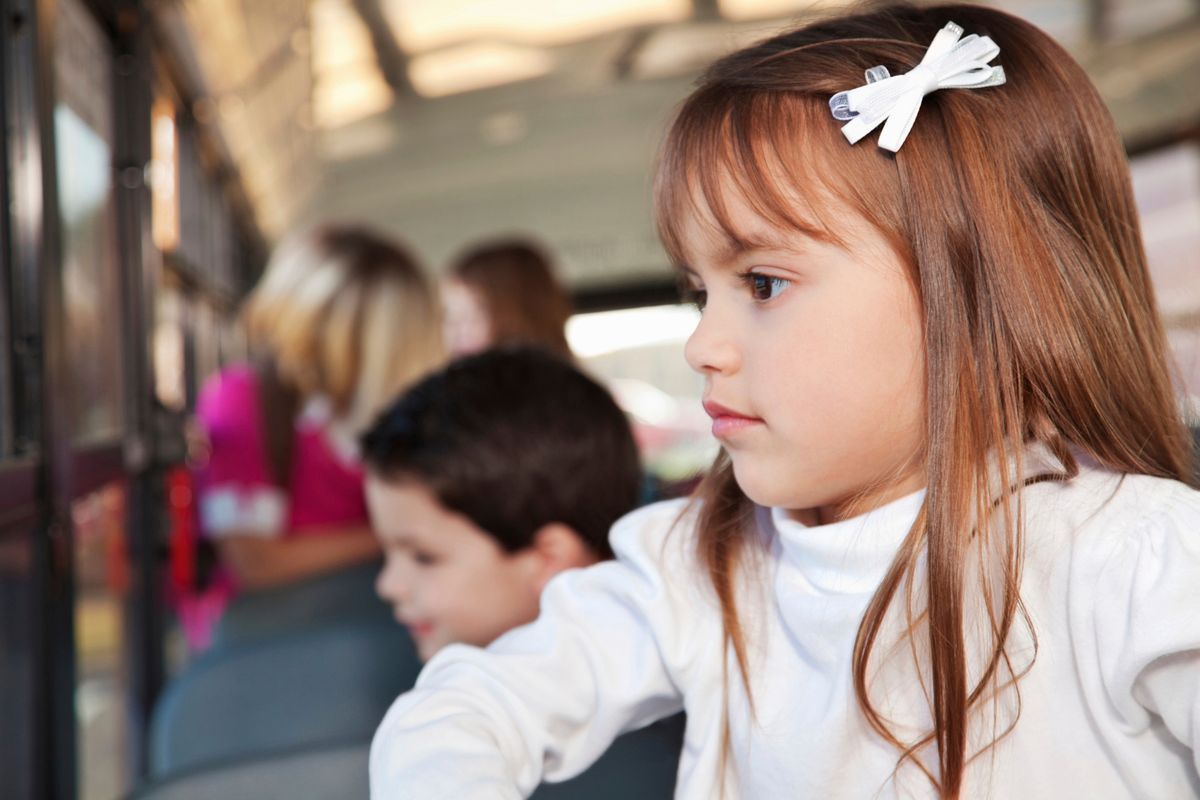 Una niña con un lazo blanco en el pelo mira por la ventana de un autobús, con un niño sentado detrás de ella.