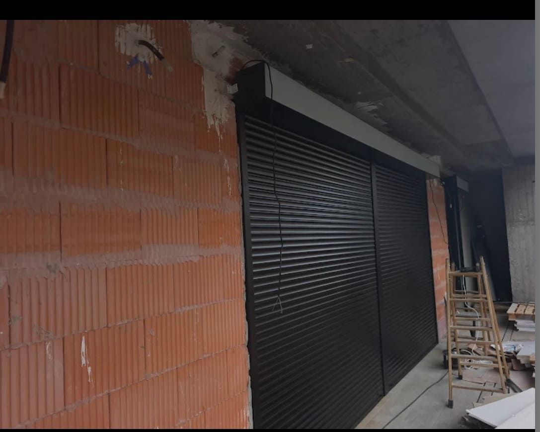 Una obra en construcción parcialmente terminada que muestra una puerta enrollable metálica negra recién instalada en una pared de ladrillo rojo.