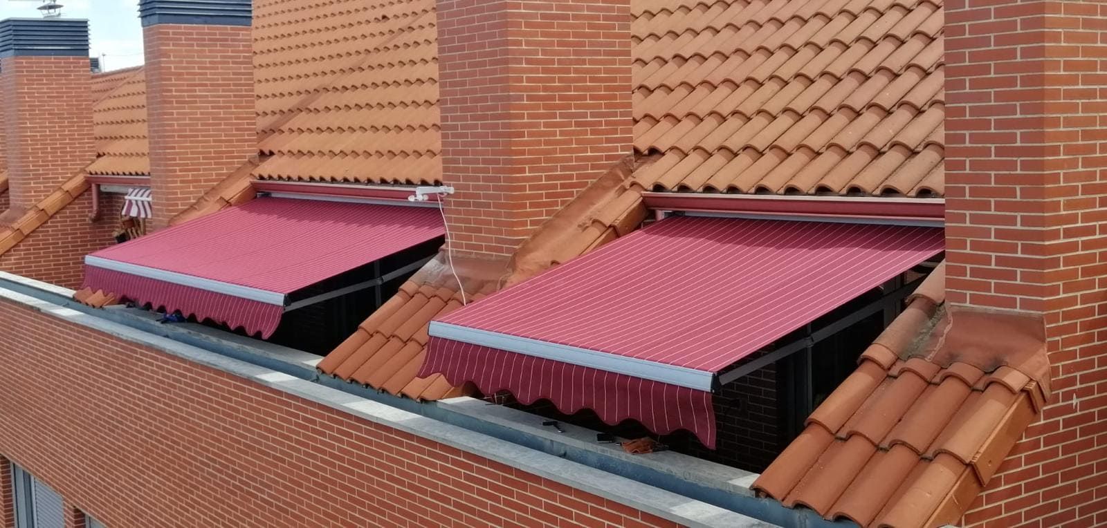 Toldos retráctiles rojos instalados bajo tejados de tejas en la fachada de un edificio de ladrillo.