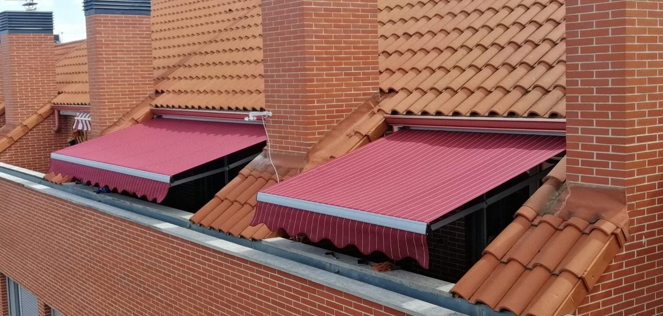 Toldos retráctiles rojos instalados bajo tejados de tejas en la fachada de un edificio de ladrillo.
