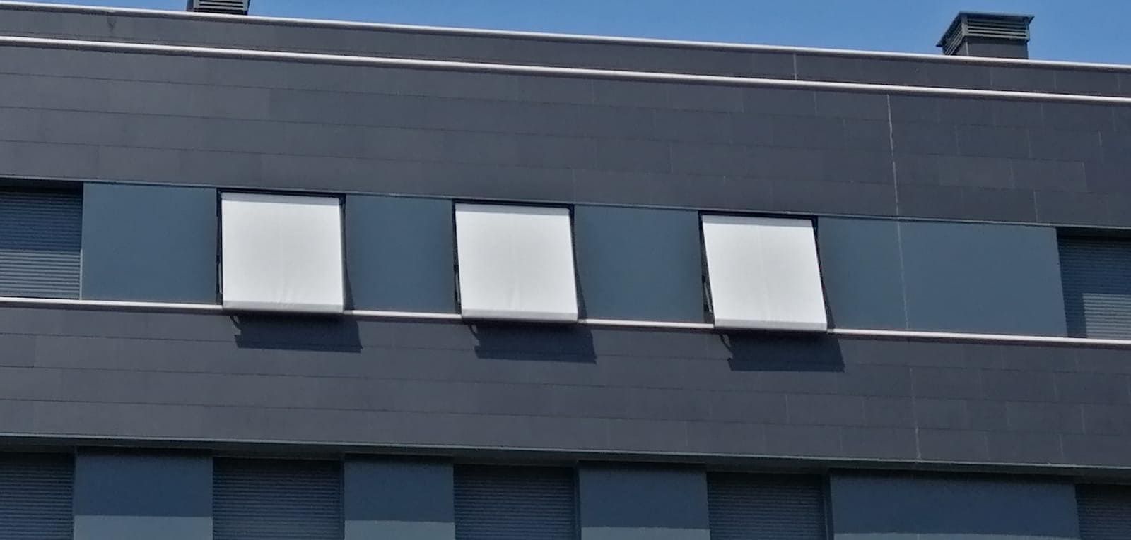 Una hilera de tres pequeñas ventanas blancas con bisagras en la parte superior, contrastando con la pared exterior revestida de paneles de color gris oscuro de un edificio moderno.