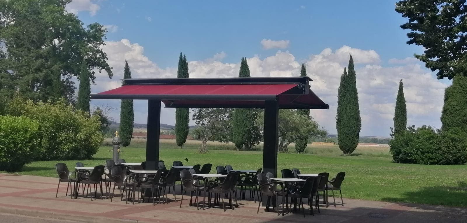Patio exterior con sillas y mesas negras bajo un pabellón de techo rojo, con vistas a un parque verde.