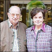 Ein älteres Ehepaar lächelt im Freien nebeneinander. Der Mann trägt eine Jacke, die Frau ein kariertes Hemd.