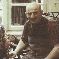 Ein Mann mit Hemd schaut in die Kamera. Er sitzt lächelnd am Fenster, umgeben von Blumen.