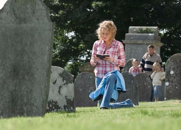Mujer arrodillada, leyendo ante una lápida en un cementerio; su familia al fondo.