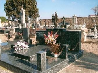 Escena de cementerio; varias lápidas de granito y marcadores de tumbas; flores y estatuas visibles; entorno soleado al aire libre.
