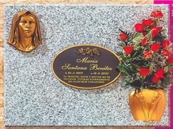 Lápida con cabeza de mujer de bronce, placa ovalada con texto y jarrón con rosas rojas.