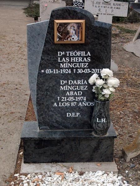 Lápida con nombres, fechas y foto. Incluye un jarrón con flores blancas.