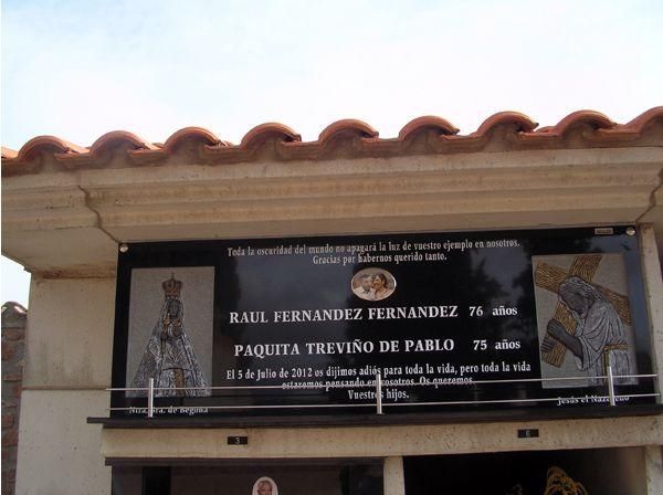 Lápida de granito negro con nombres, fechas e imágenes religiosas bajo techo.