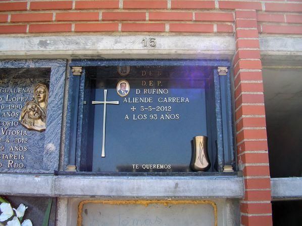 Un monumento con una cruz, una foto y un texto sobre un fondo azul, en una pared de ladrillos.