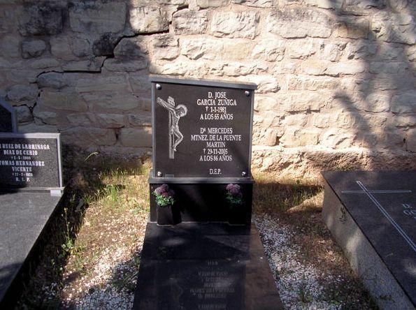 Lápida de granito negro con cruz e inscripción, situada en un cementerio con fondo de muro de piedra.