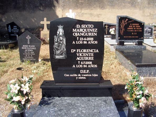 Una lápida de granito negro con nombres y fechas, flores en el frente y otras lápidas en un cementerio.