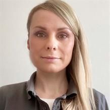 Eine Frau mit langen blonden Haaren trägt ein graues Hemd und lächelt in die Kamera.
