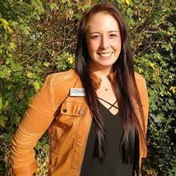 Eine Frau mit einer braunen Jacke und einem schwarzen Hemd steht vor einem Busch.