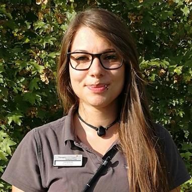 Eine Frau mit Brille und Namensschild steht vor einem Baum.