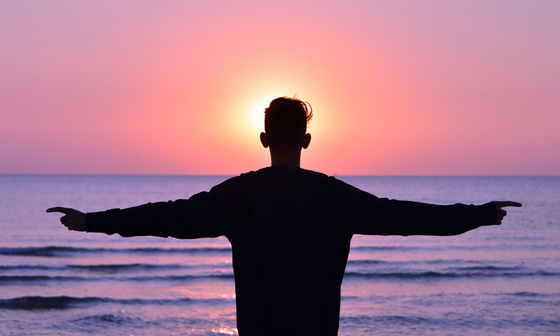 Un hombre está de pie en la playa con los brazos extendidos al atardecer.