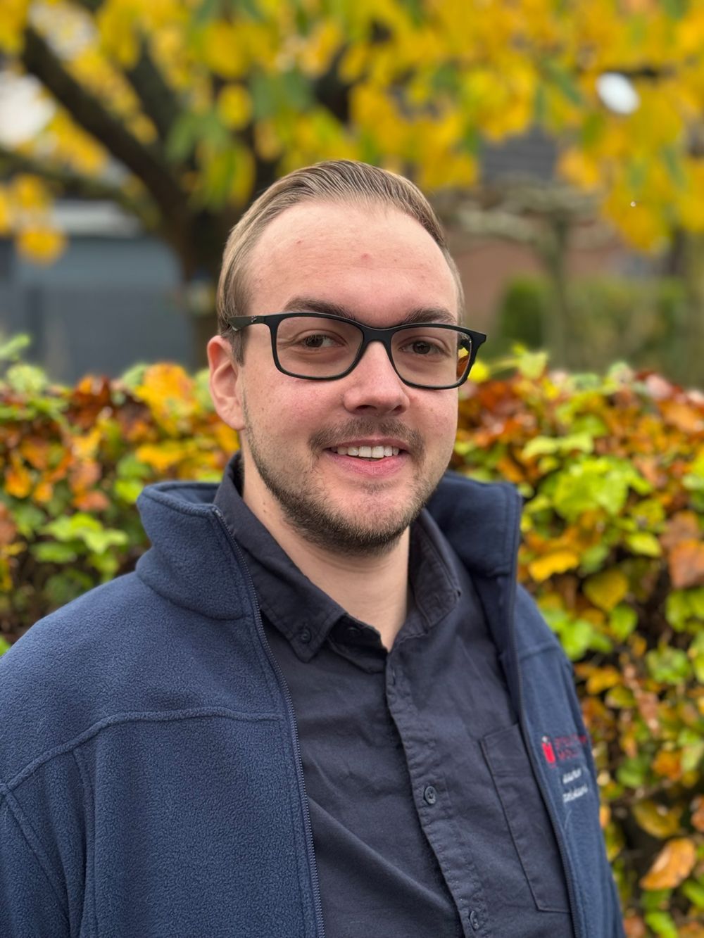Ein Mann mit Brille lächelt, er trägt eine blaue Fleecejacke und ein Hemd mit Kragen, er steht draußen im Herbstlaub.