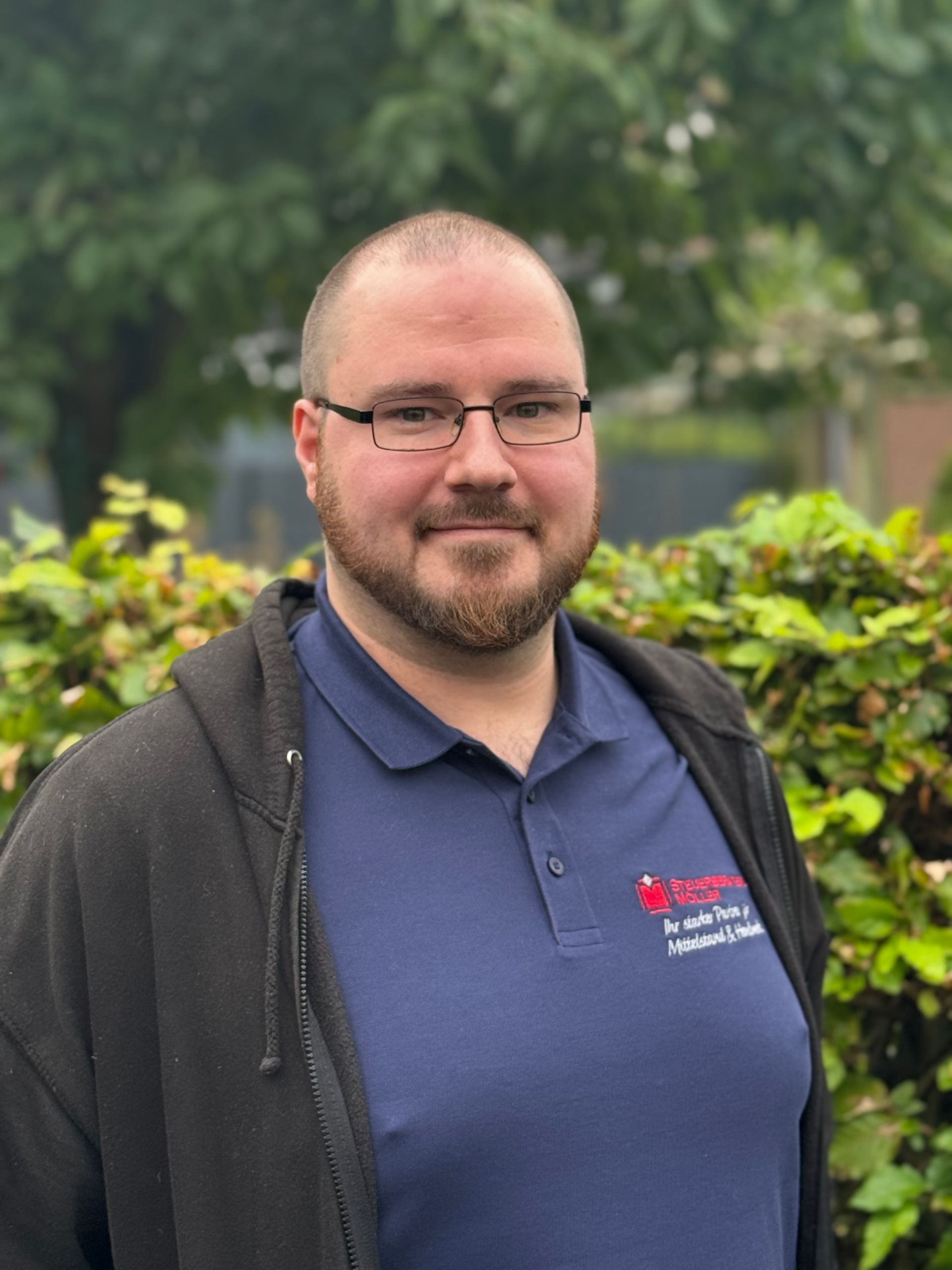 Ein Mann mit Bart lächelt vor einem Baum mit weißen Blüten.