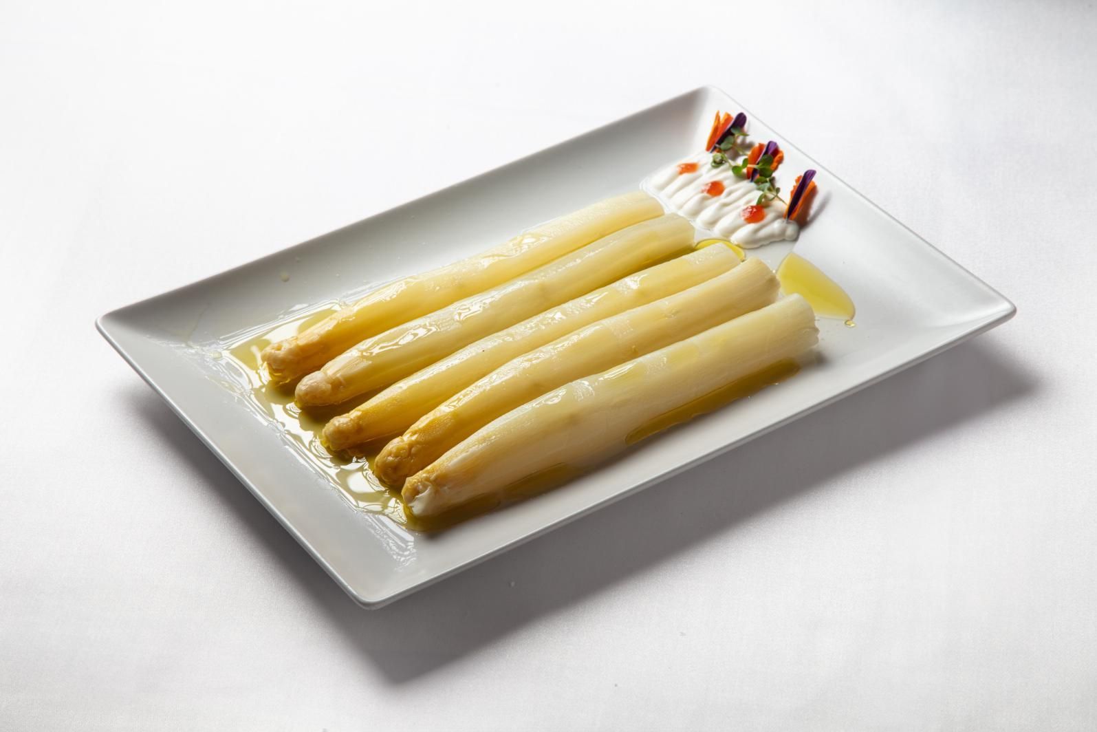 Cuatro espárragos blancos en un plato rectangular con guarnición de salsa.