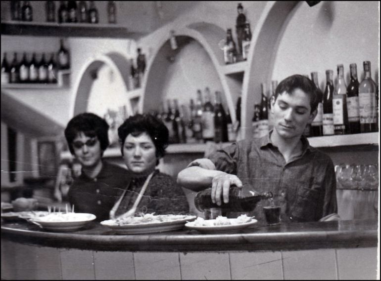 Tres personas están sentadas en la barra de un bar, con botellas en los estantes, y un hombre sirve comida en una cafetería.