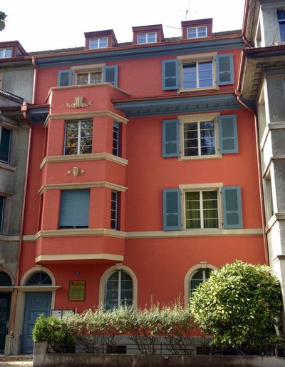 Korallenfarbenes Gebäude mit hellblauen Fensterläden und Zierelementen, Erkerfenster, Bogentür und kleinem Gebüsch.