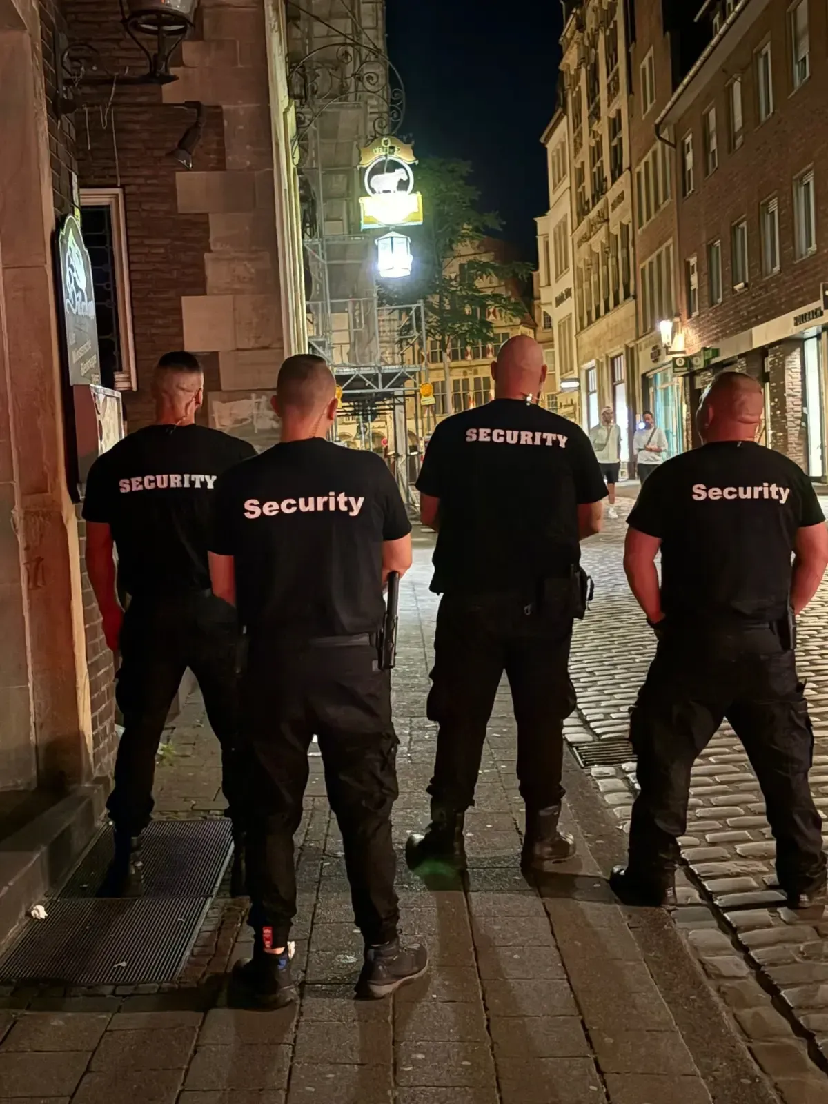 Vier Sicherheitsleute in schwarzen Uniformen stehen nachts auf einer Kopfsteinpflasterstraße und blicken mit dem Rücken zur Kamera.