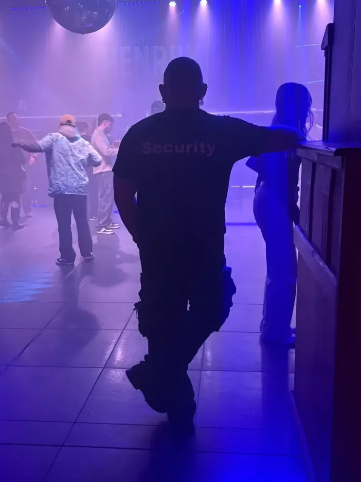Die Silhouette eines Wachmanns steht in einem dunklen, blau beleuchteten Nachtclub, im Hintergrund tanzen die Gäste.