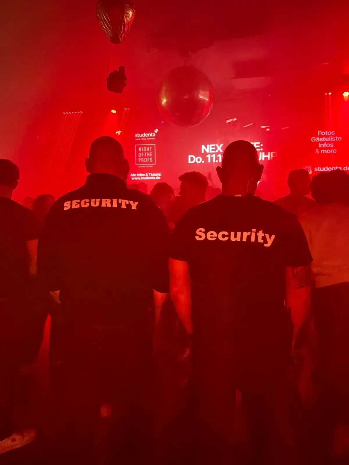 Zwei Sicherheitsleute in schwarzen Hemden stehen in einem schwach beleuchteten, rotgetönten Club und blicken auf eine Menschenmenge und eine Bühne mit einer Discokugel.